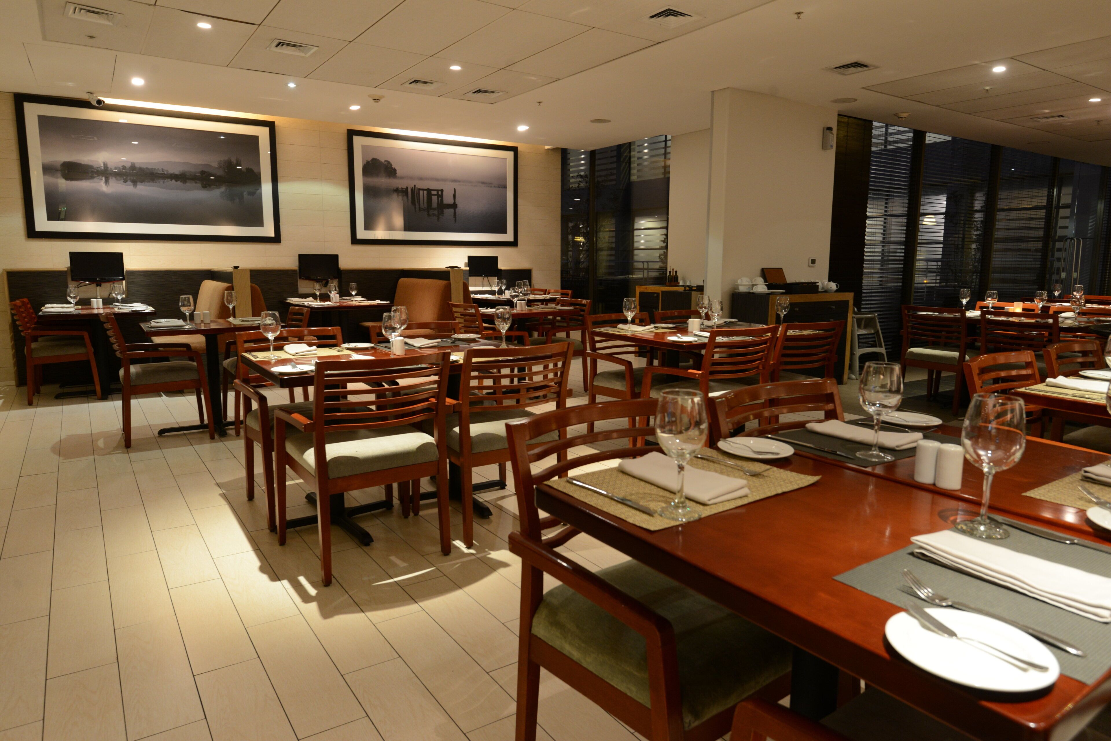A restaurant with multiple dining tables and chairs, framed artwork on the walls, and a view of the city through large windows.