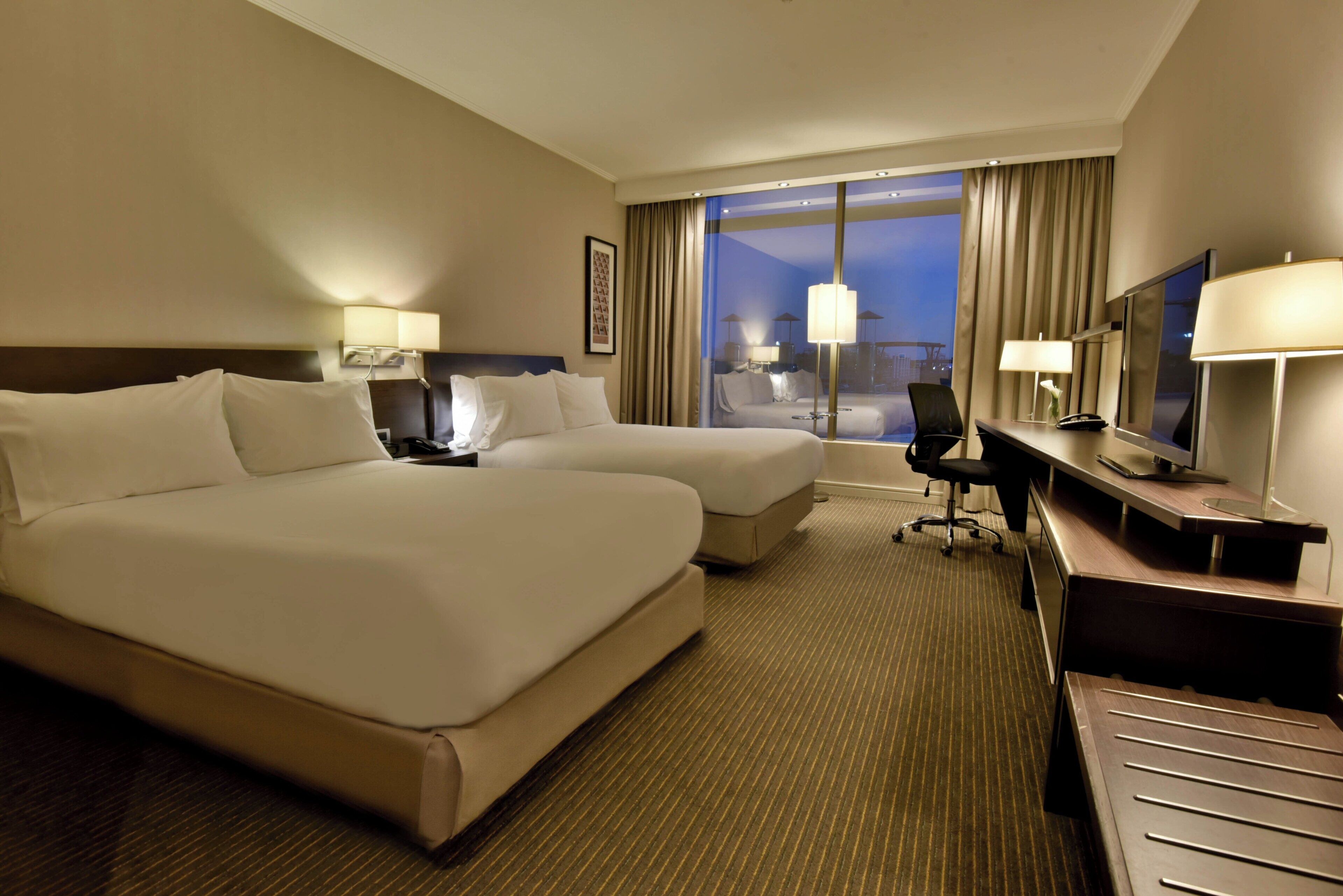 A hotel room with two beds, a desk, a chair, a TV, and a window with curtains.