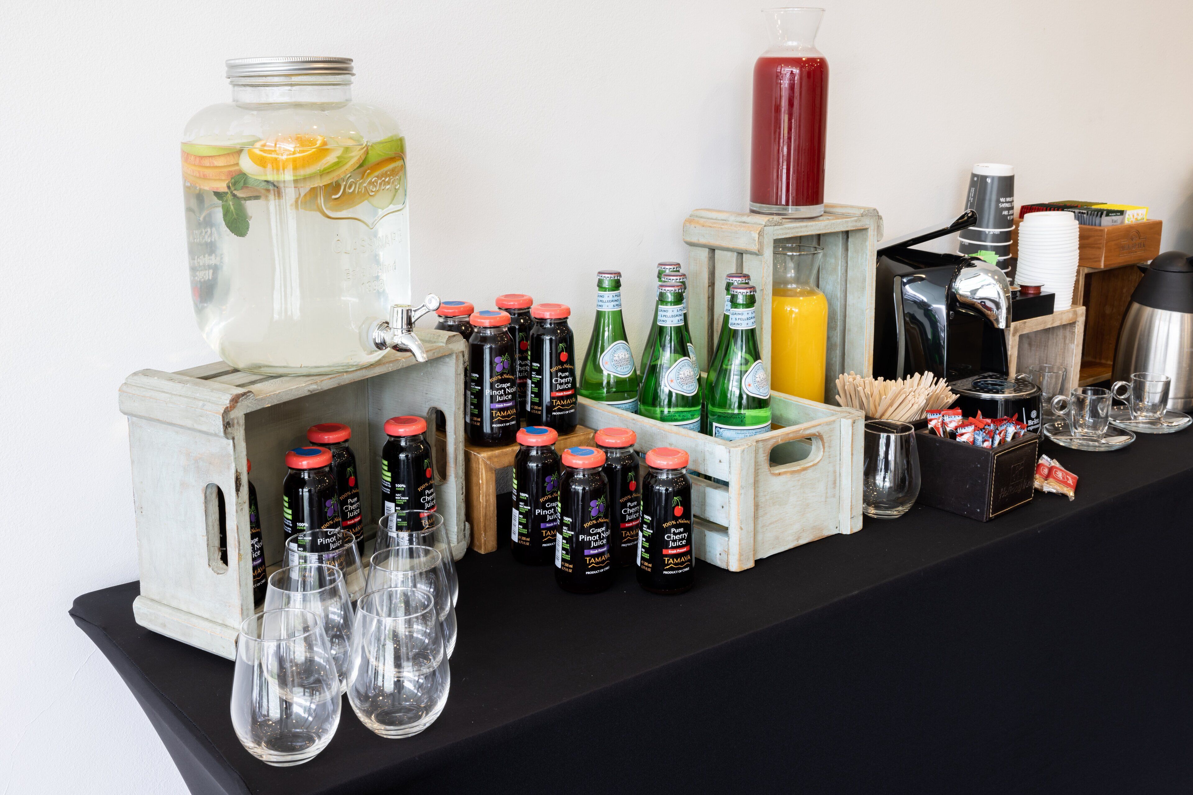 A bar setup with a large pitcher, multiple bottles of beverages, glasses, and a coffee maker.