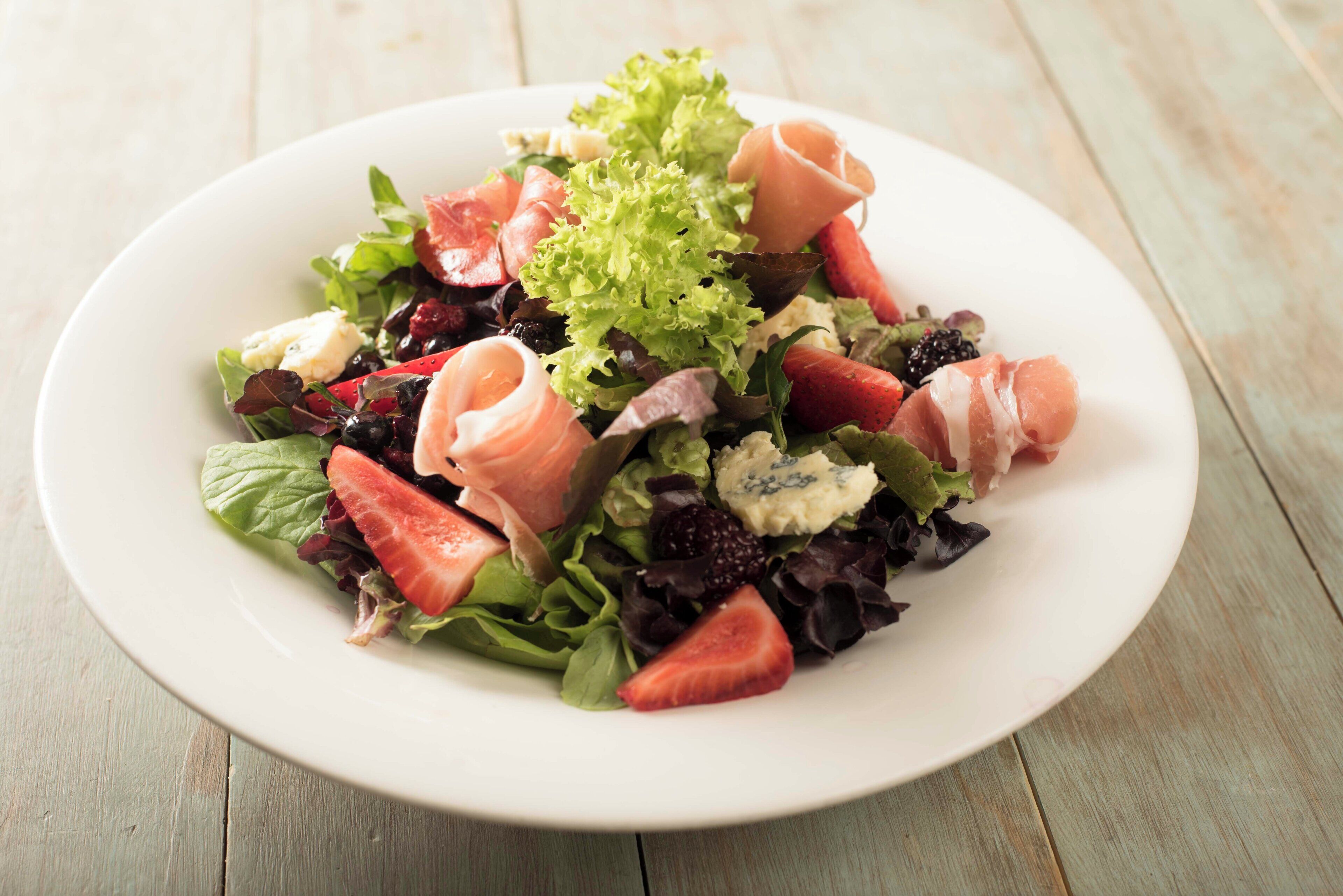 Uma salada de folhas verdes mistas com morangos, framboesas e fatias de salmão defumado, sobre um prato branco.