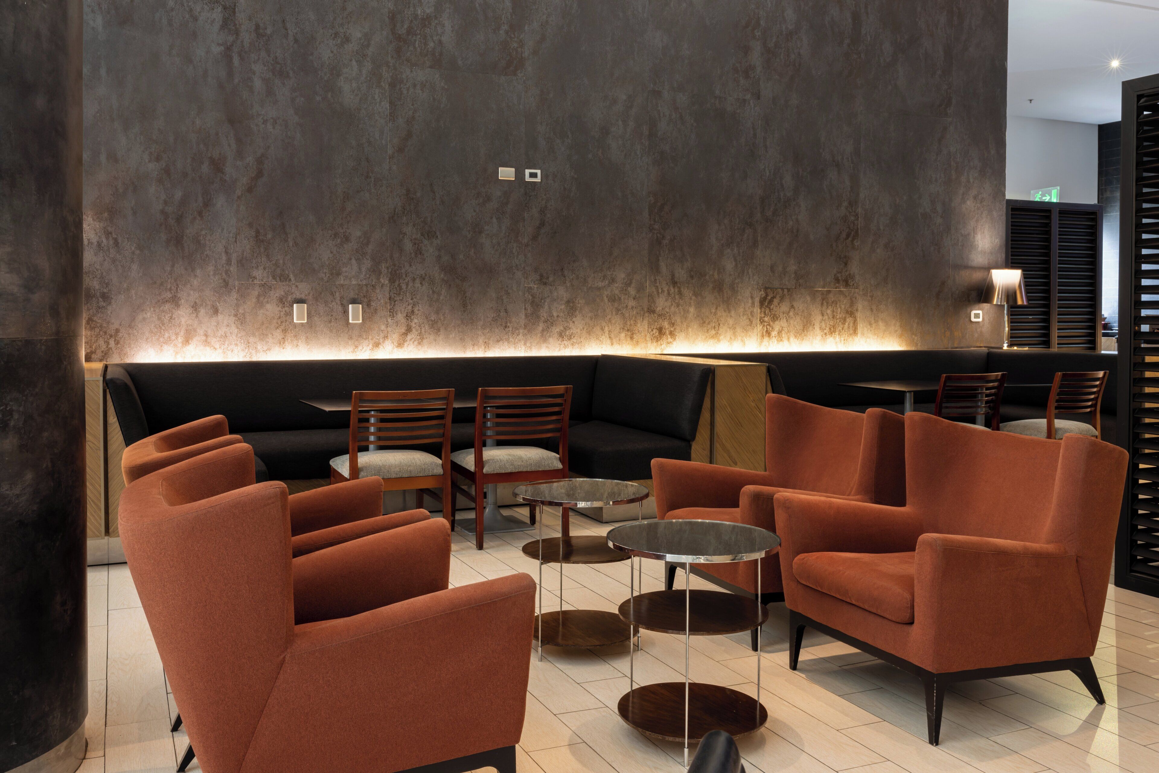 Un área de descanso moderna con sofás, sillones y mesitas, que destaca por una pared con textura e iluminación ambiental.