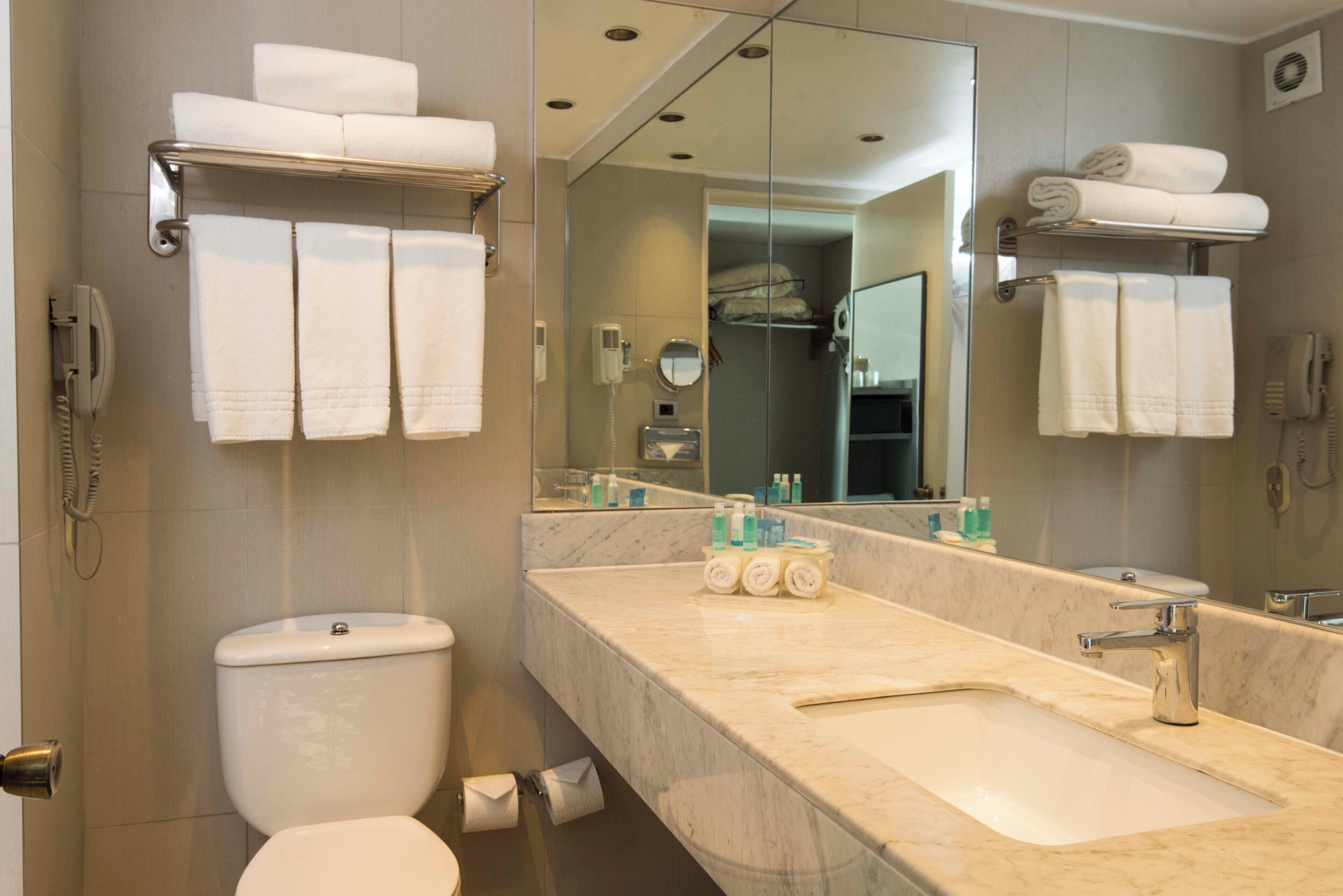 Een badkamer met toilet, wastafel en spiegel. Handdoeken liggen op een rek en er hangt een telefoon aan de muur.