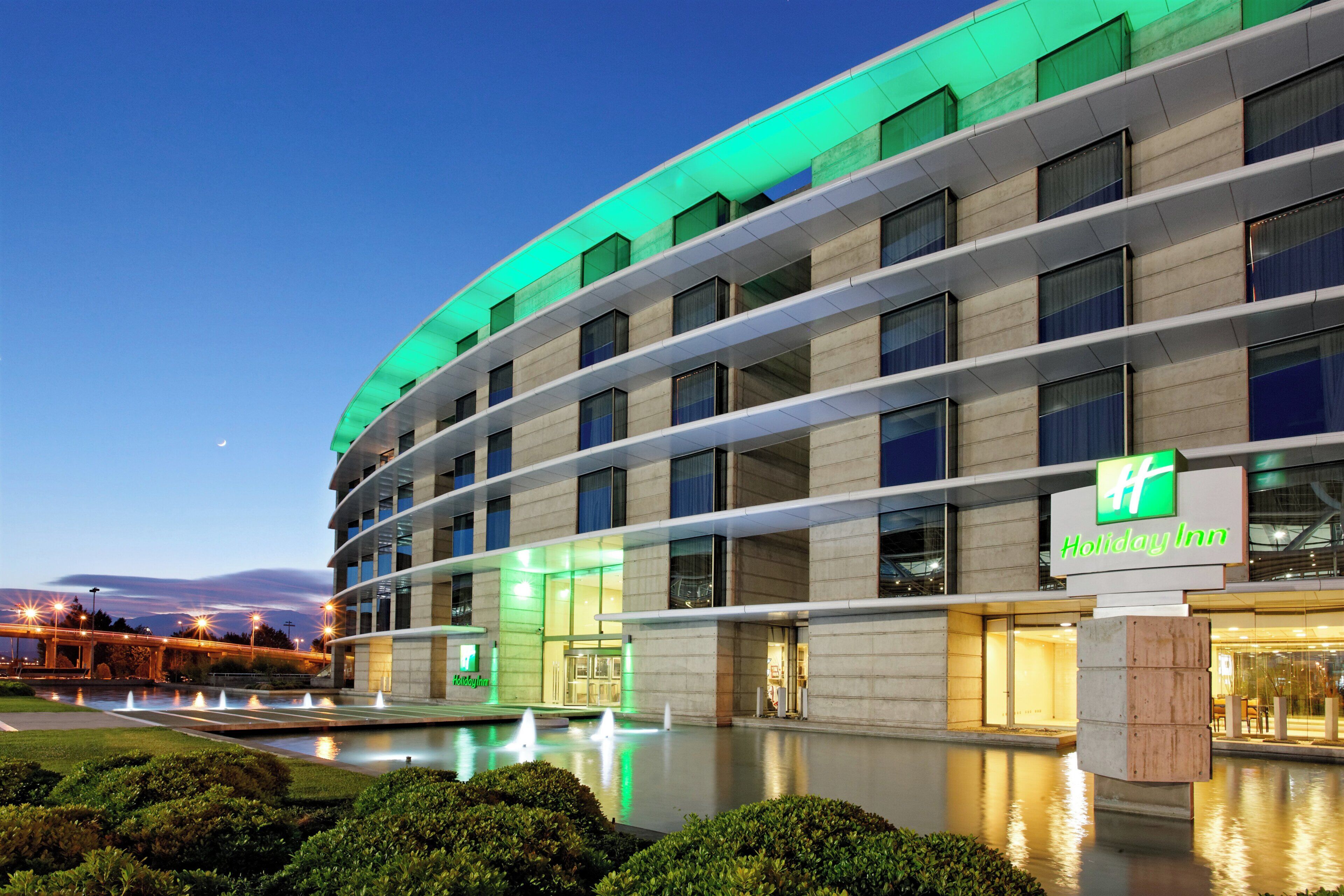 Un hotel Holiday Inn con diseño moderno, que cuenta con una gran fuente en primer plano, varios pisos y un llamativo letrero verde.