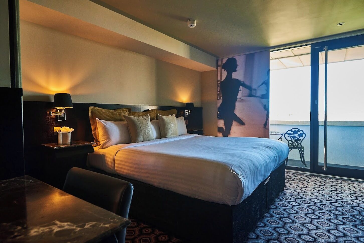 Ein Hotelzimmer mit Bett, Nachttisch mit Lampe, Stuhl, Tisch und einem Balkon mit Blick.