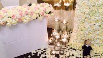 A wedding reception area with a table covered in white and pink flowers, candle-lit glass vases, and a teddy bear on the floor.