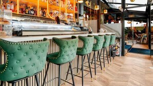 Zona de bar con taburetes tapizados de verde, una barra con botellas y estantes con diversos objetos.