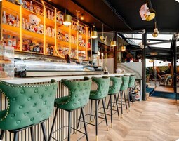 Un espace bar avec des tabourets de bar à assise rembourrée de couleur verte, un comptoir de bar orné de bouteilles et des étagères remplies de divers objets.