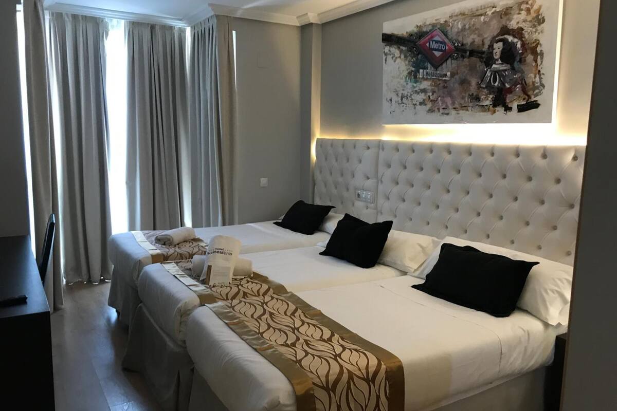 Habitación de hotel con dos camas, un cabecero grande tapizado, un cuadro en la pared y vistas al exterior a través de la ventana.