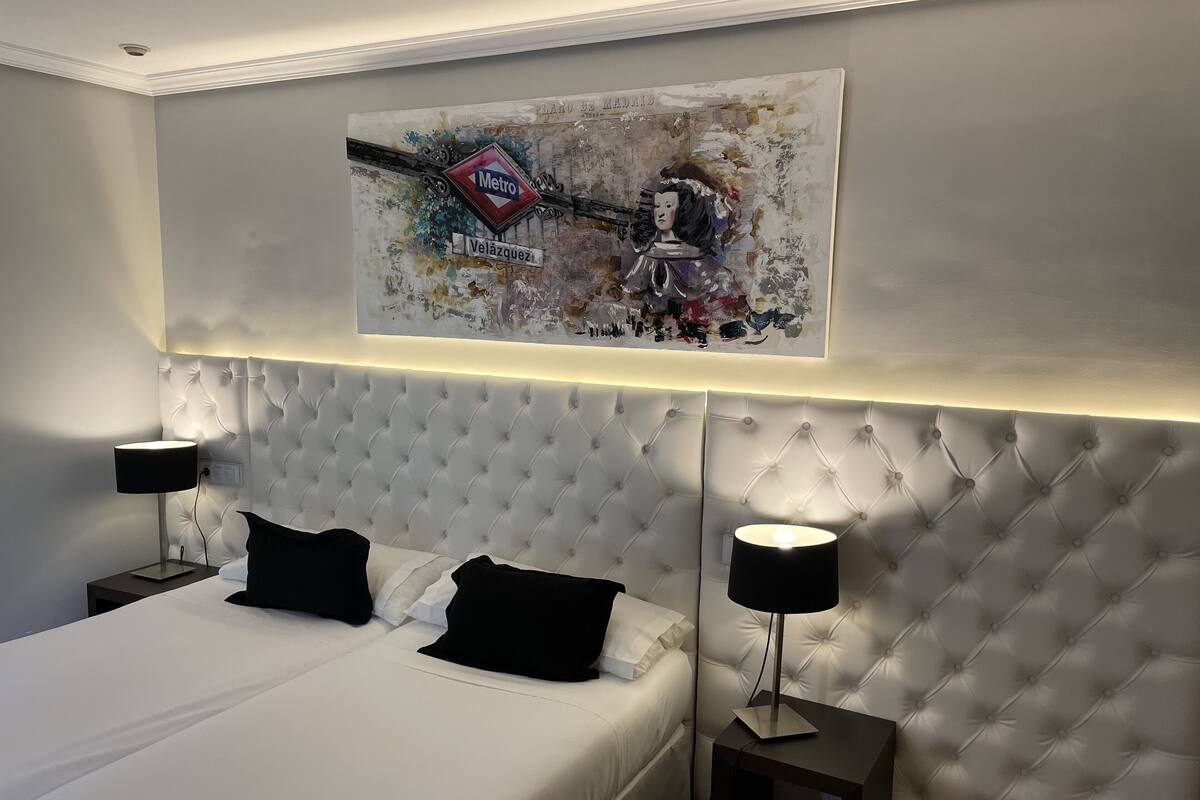 Un dormitorio moderno con un cabecero capitonado, una cama grande con ropa blanca, dos mesitas de noche con lámparas, y una obra de arte en la pared que muestra una escena de metro.