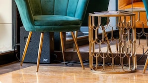 Una butaca tapizada en color verde agua con base dorada, una mesa redonda con base dorada y un vidrio que refleja follaje exterior.