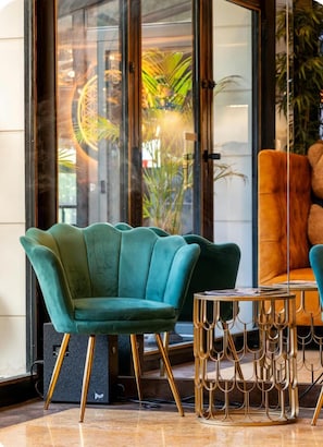 A teal upholstered chair with a gold base, a round table with a gold base, and a glass door reflecting outdoor greenery. - Hotel Suites Feria de Madrid (Madrid)