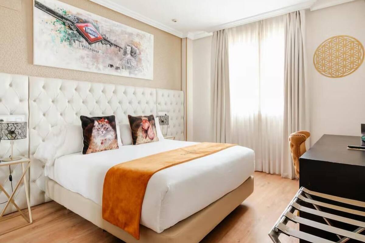 Una habitación de hotel con un cabecero capitonado, una cama con edredón blanco y manta naranja, un escritorio con silla y un gran ventanal con cortinas.