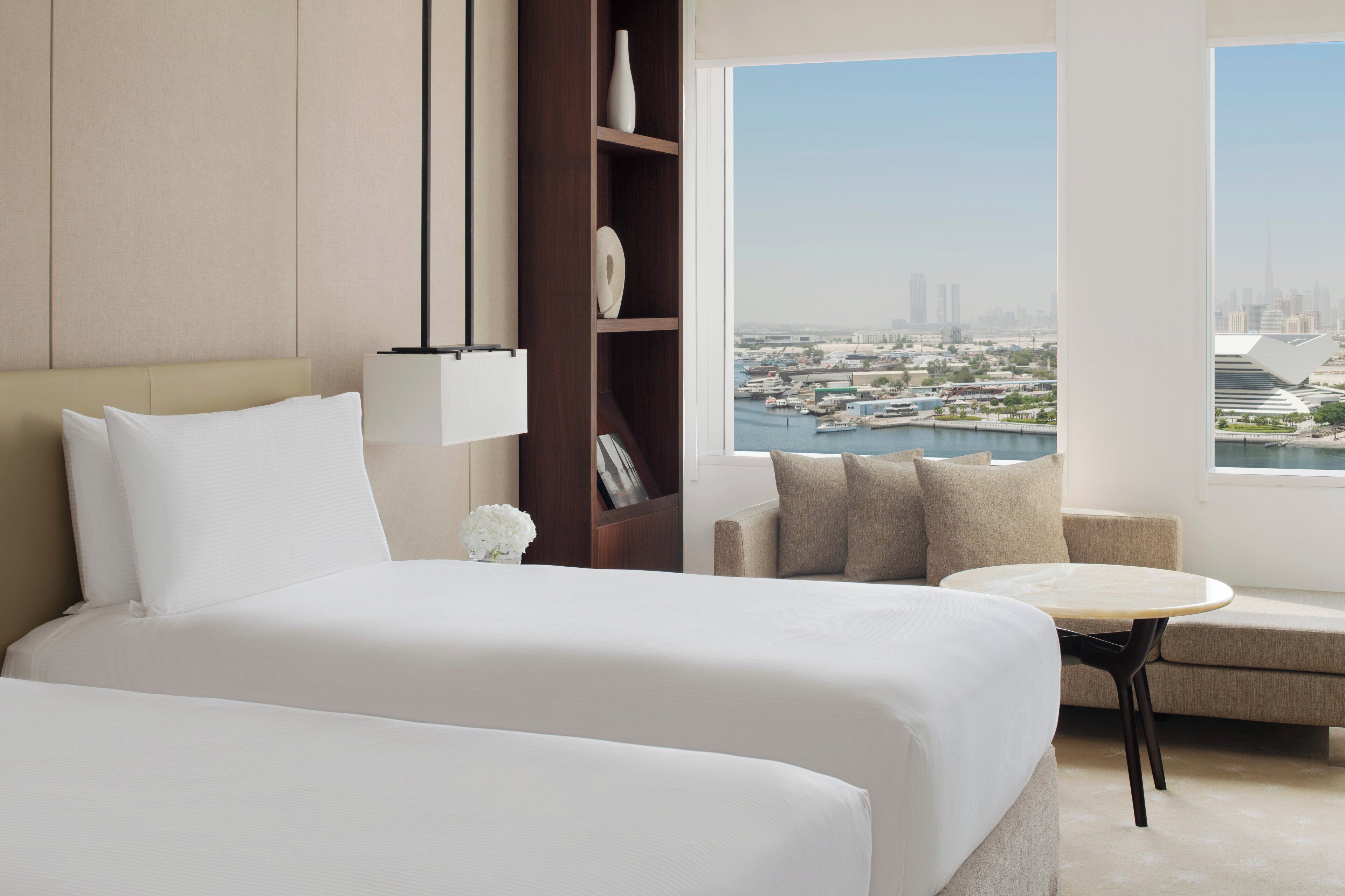 A hotel room with a bed, a sofa, a small table, a bookshelf, and a view of a cityscape.