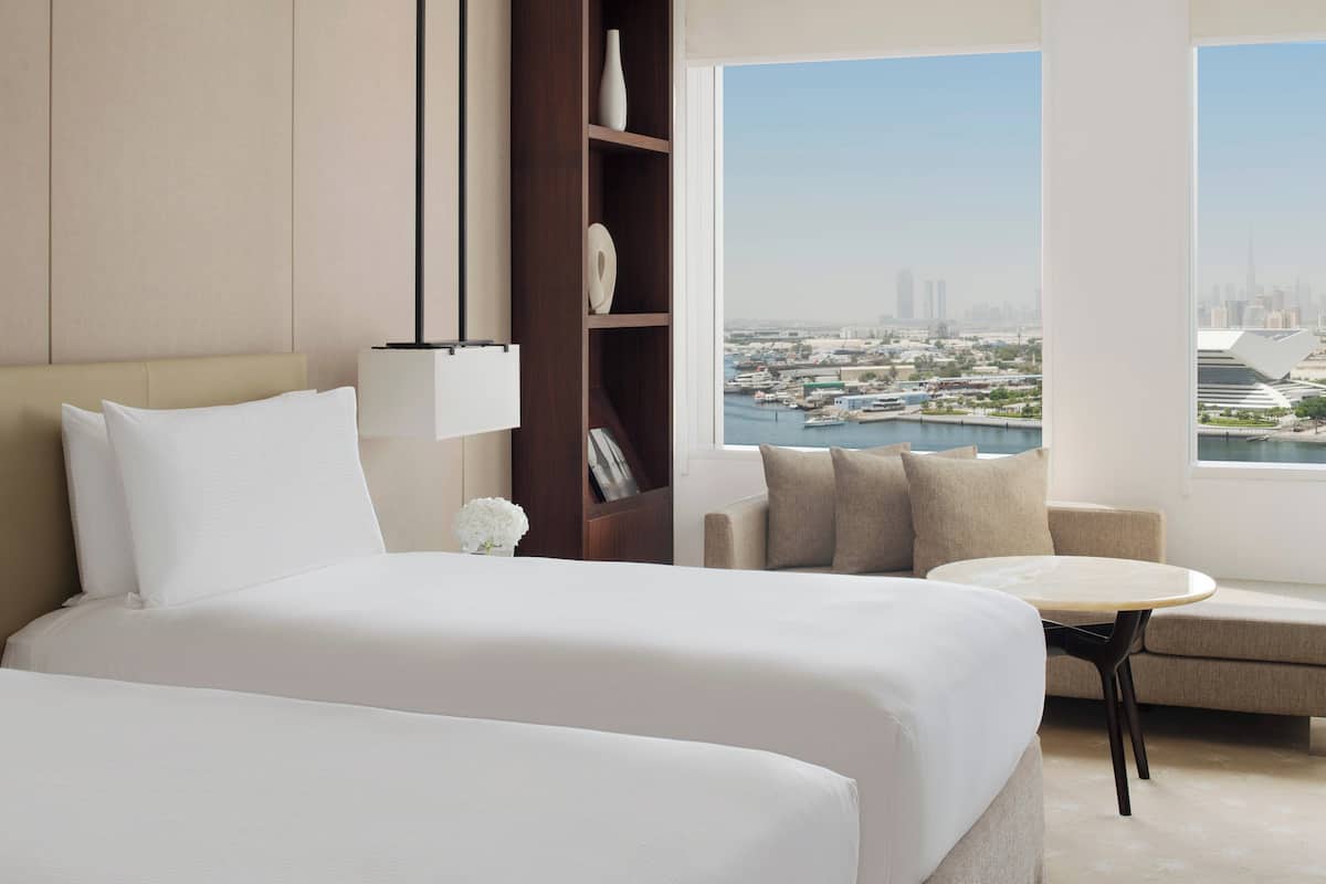 Habitación de hotel con cama, sofá, una mesita, una estantería y vista a la ciudad.
