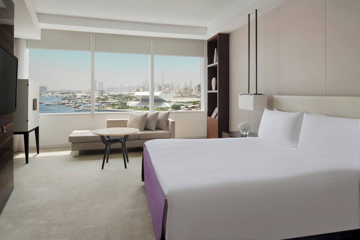 Habitación de hotel moderna con una cama grande, un sofá, una mesita y vistas a un paisaje urbano a través de amplios ventanales.