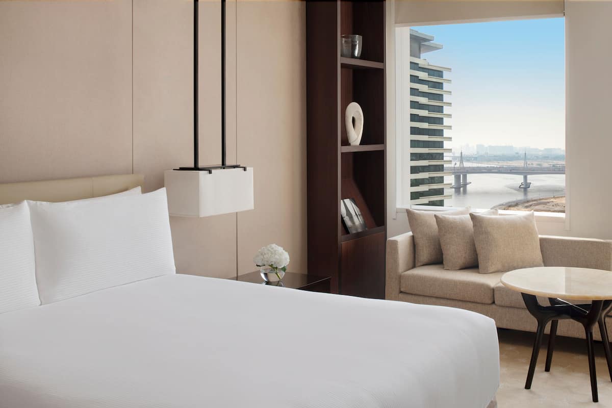 Habitación de hotel con una cama grande, un sofá, una mesita, una estantería y vistas a un puente y al paisaje urbano.