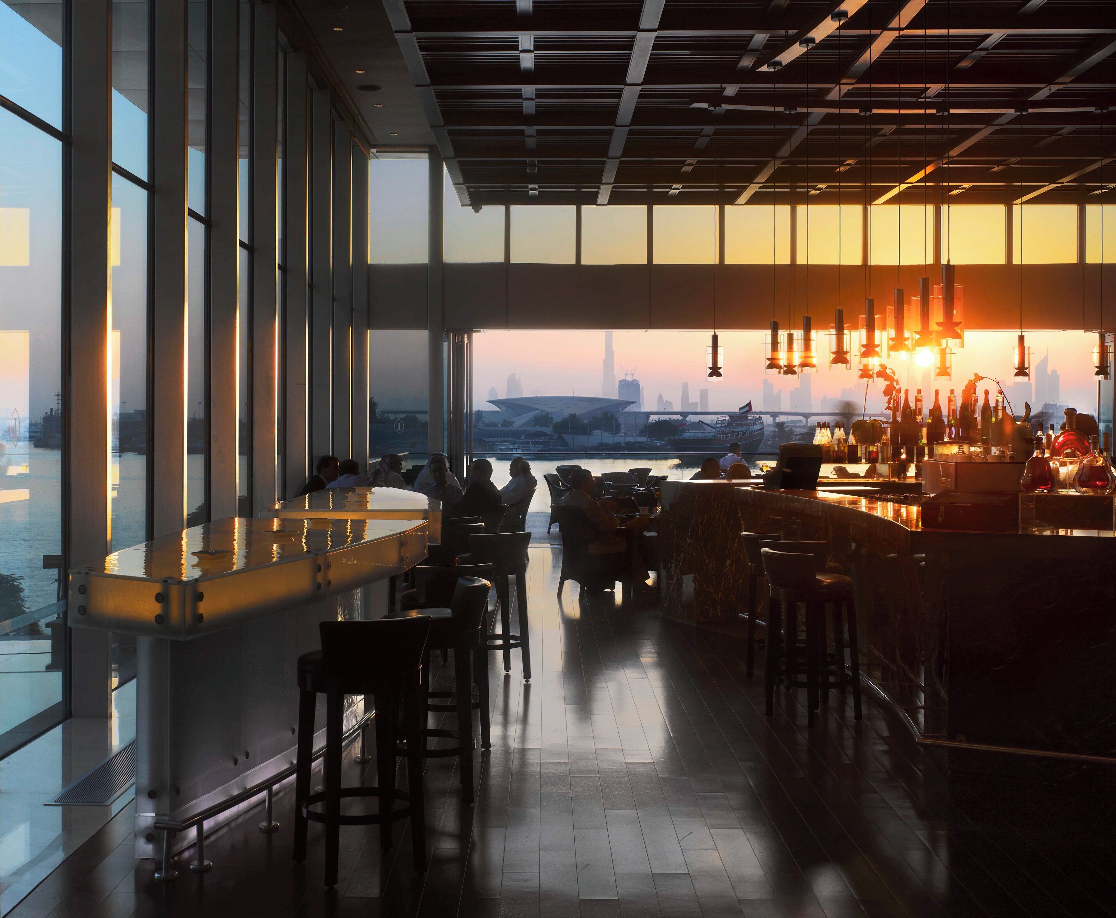 A bar with a view of a city skyline at sunset, large windows, and modern interior design.