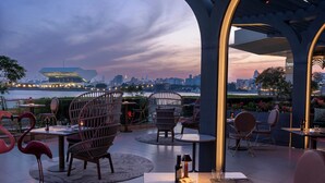 Un espacio de comedor al aire libre con mesas redondas y sillas de mimbre, con vistas a un horizonte urbano y una masa de agua.