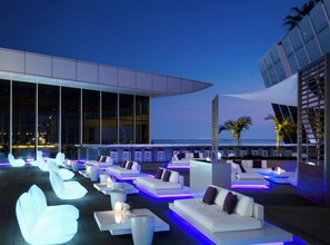 Un salon sur le toit, avec des canapés blancs et des tabourets de bar, une table de billard et une vue sur les toits de la ville au crépuscule.