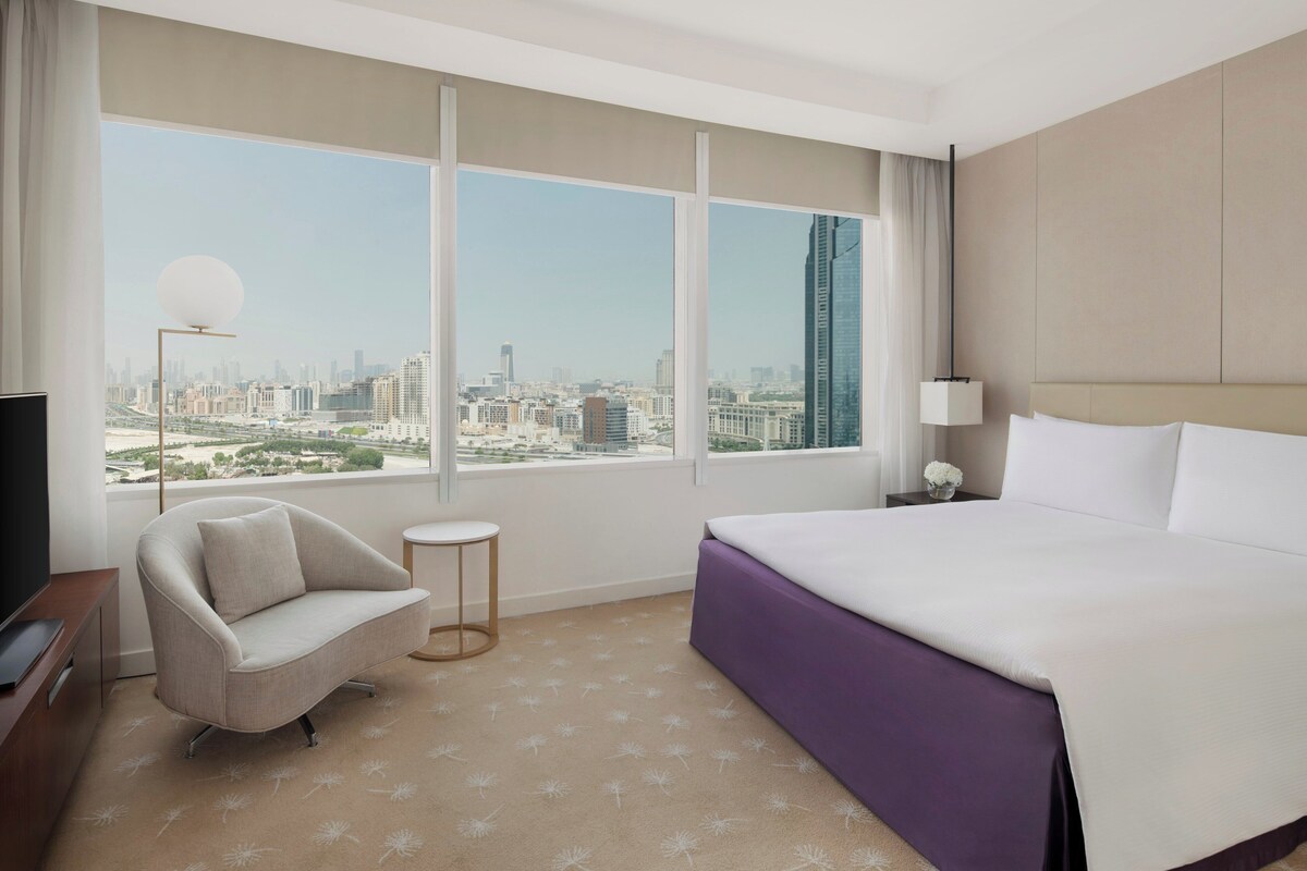 Ein Hotelzimmer mit einem großen Bett, einem Sofa, einem kleinen Beistelltisch, einem Fernseher und einem Fenster mit Blick auf die Stadt.
