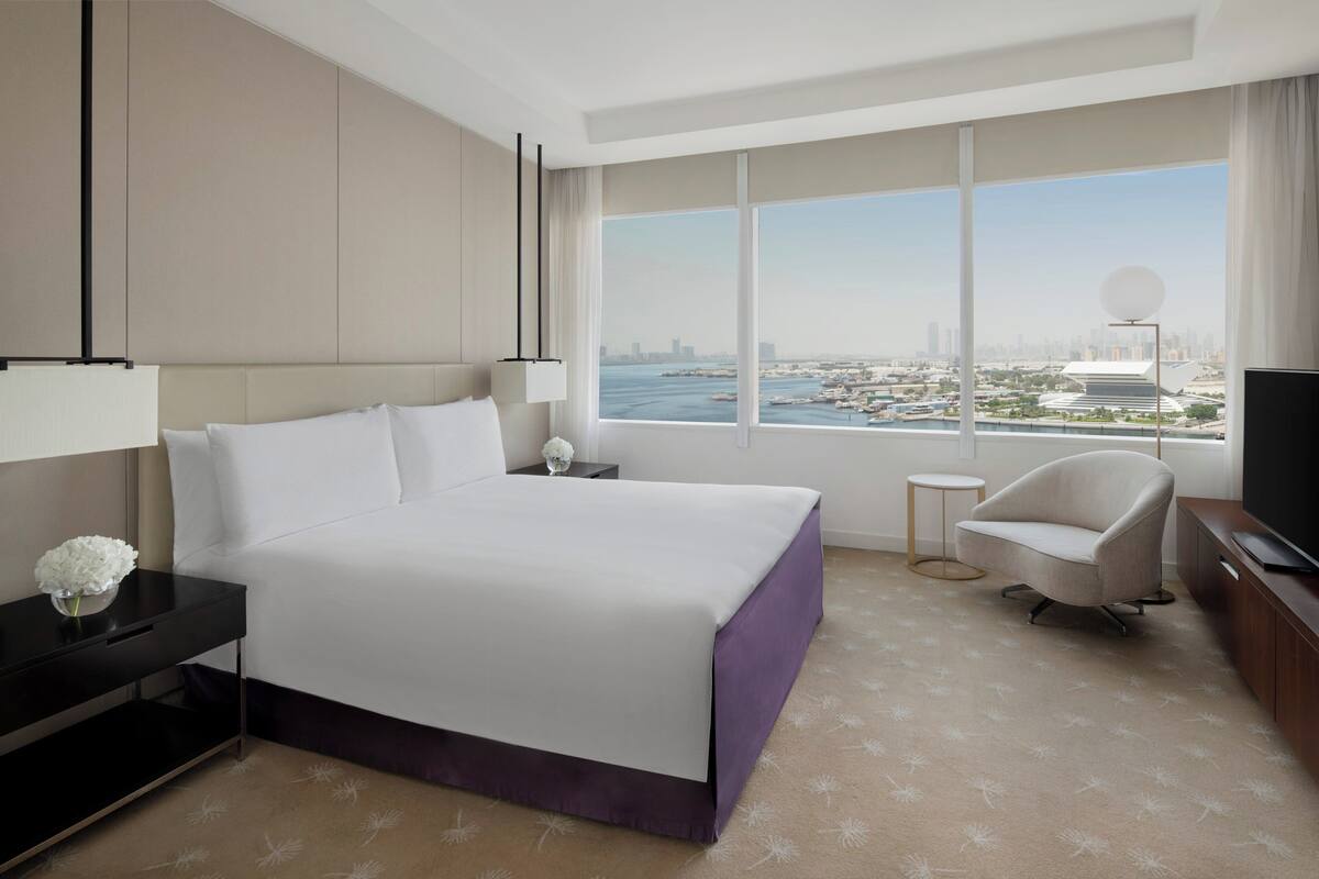 Una habitación de hotel moderna con una cama grande, televisión, una silla y vistas a la ciudad a través de amplios ventanales.