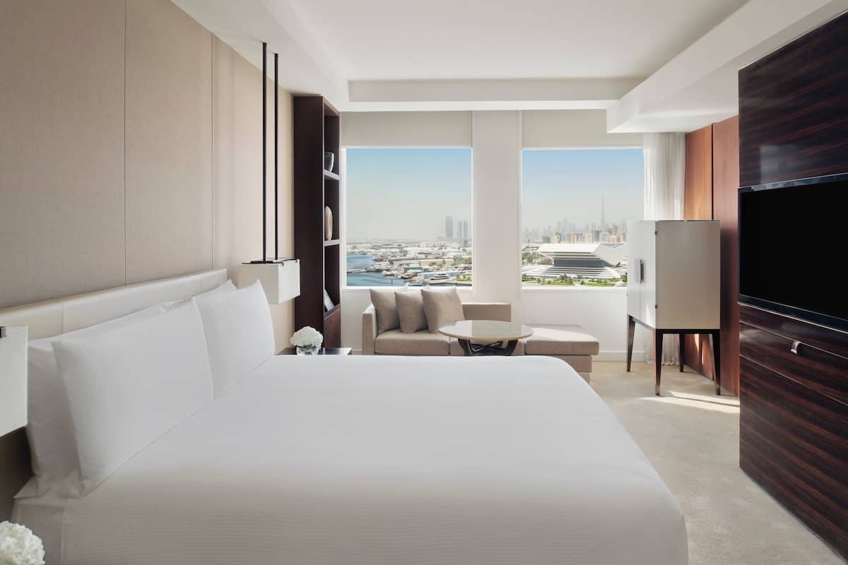 Una habitación de hotel moderna con una cama grande, un sofá, una mesita, un televisor y vistas a un paisaje urbano.