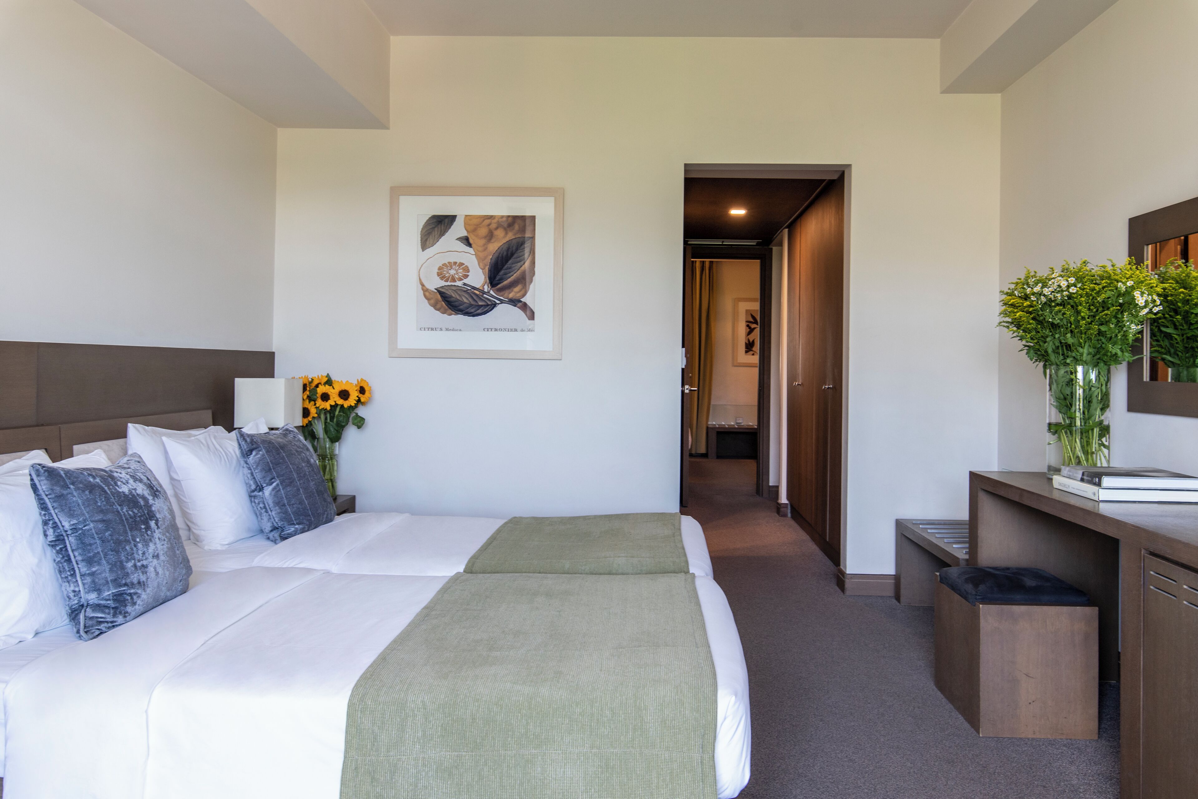 A hotel room with a bed, a desk, a chair, a vase with flowers, and a framed botanical illustration on the wall.
