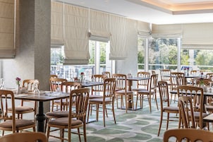 A spacious dining area with multiple tables and chairs, large windows with curtains, and a view of greenery outside.