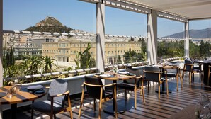 Un restaurant situé sur le toit d’un immeuble, avec des tables et des chaises, de grandes fenêtres offrant une vue sur le paysage urbain et une colline.