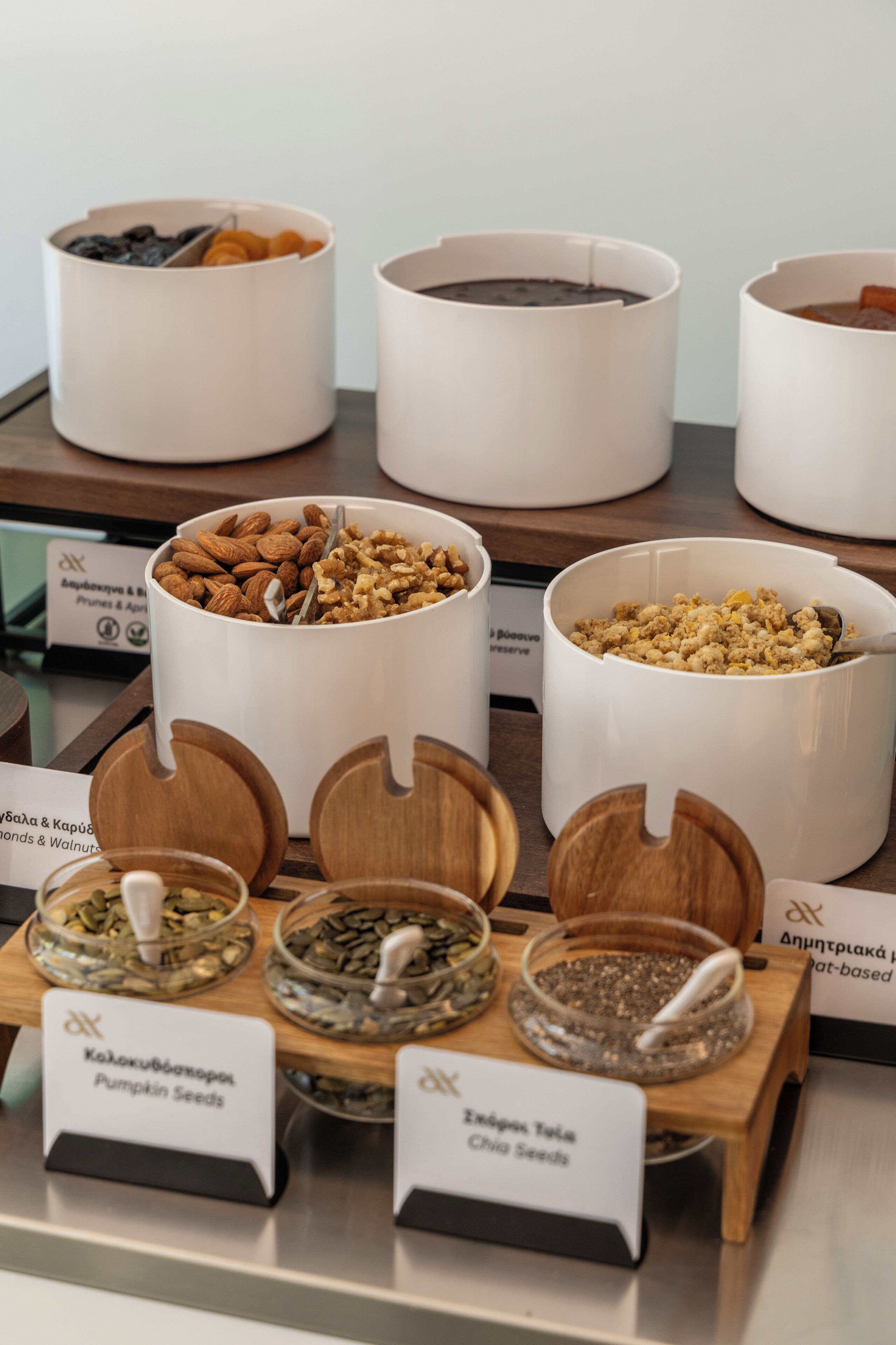 A selection of nuts and seeds in white containers with wooden lids, some labeled with their names.