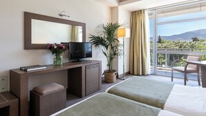 Une chambre d’hôtel avec un lit, un bureau avec une télévision, une plante et un balcon avec vue.