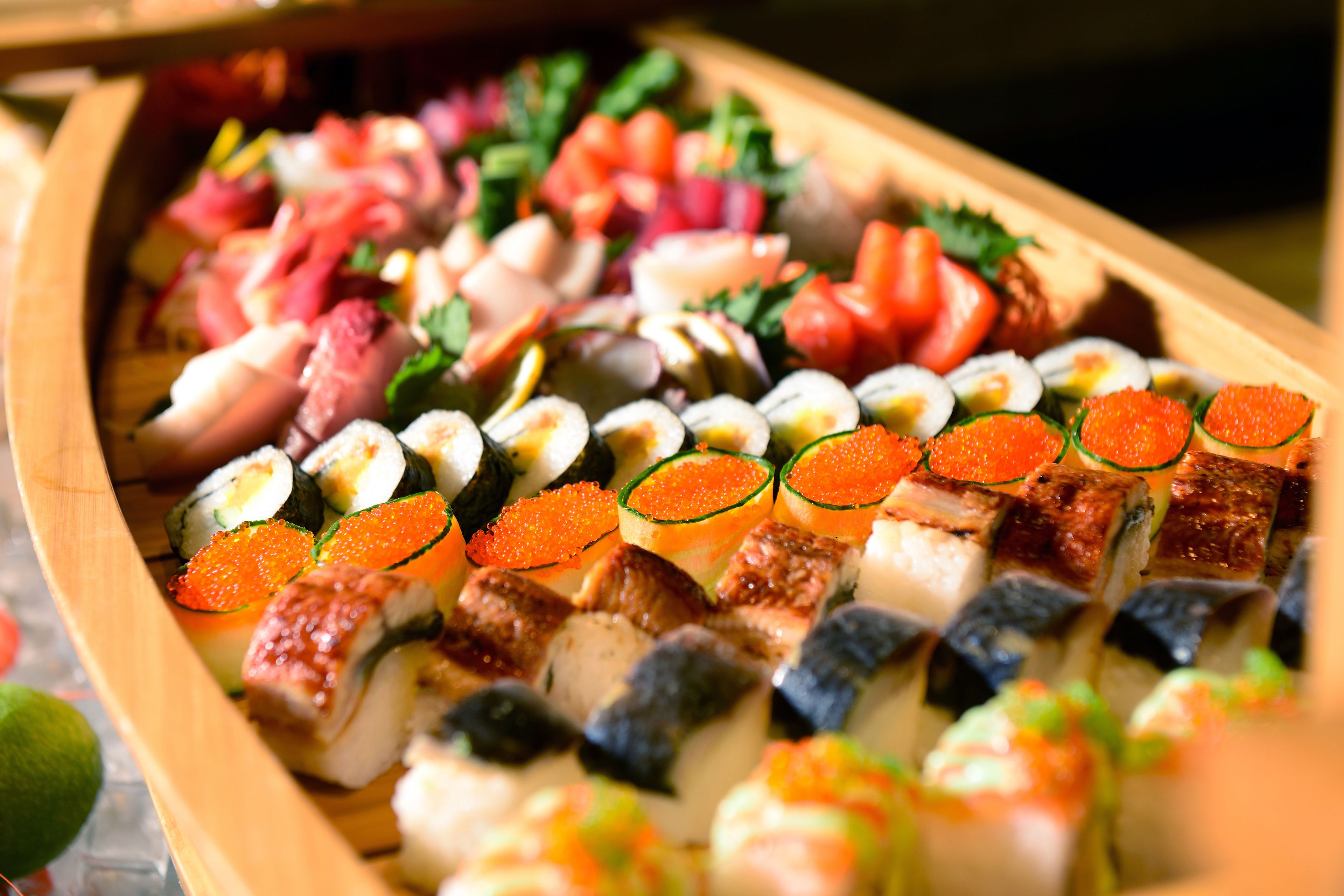 A wooden tray filled with various sushi pieces, including rolls and nigiri, garnished with vegetables and fish roe.