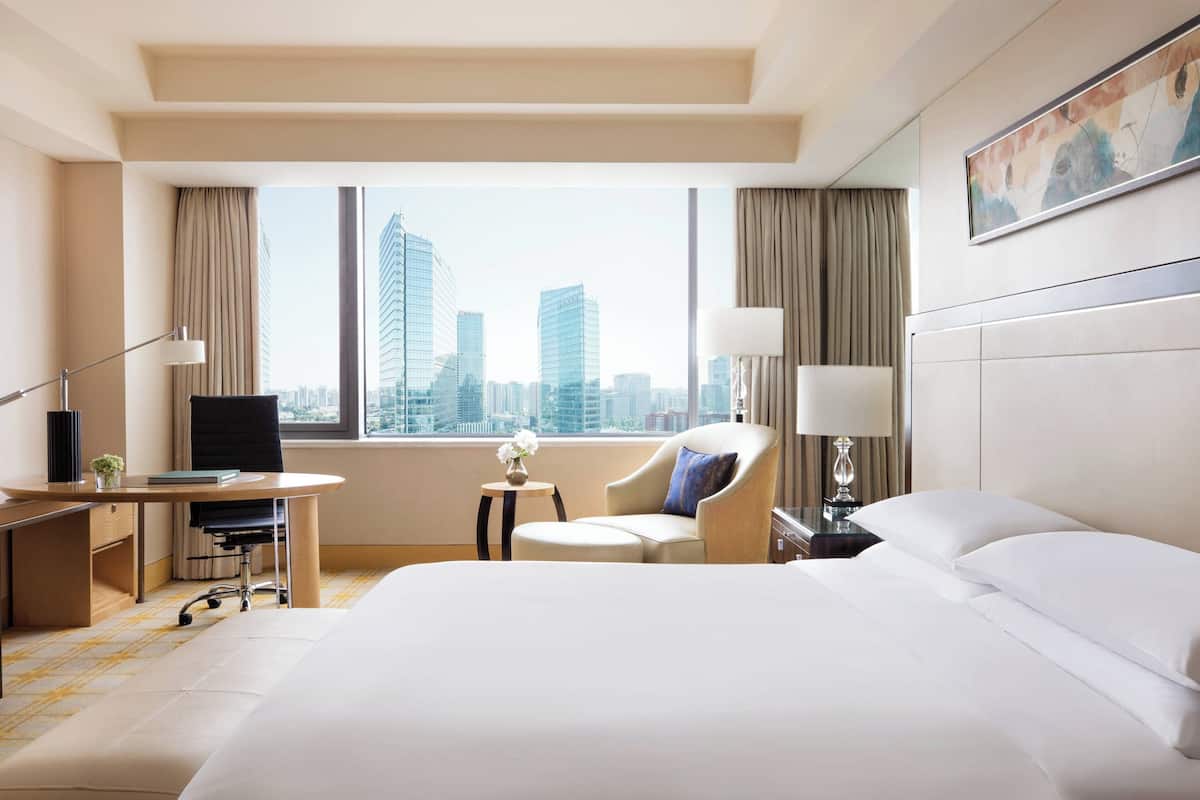 A hotel room with a large bed, a desk with a chair, a small table with a lamp, and a view of tall buildings.