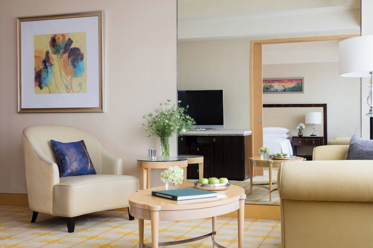 A hotel room with a beige sofa, a round wooden coffee table with a book and a vase of flowers, a television, and a bed in the background.