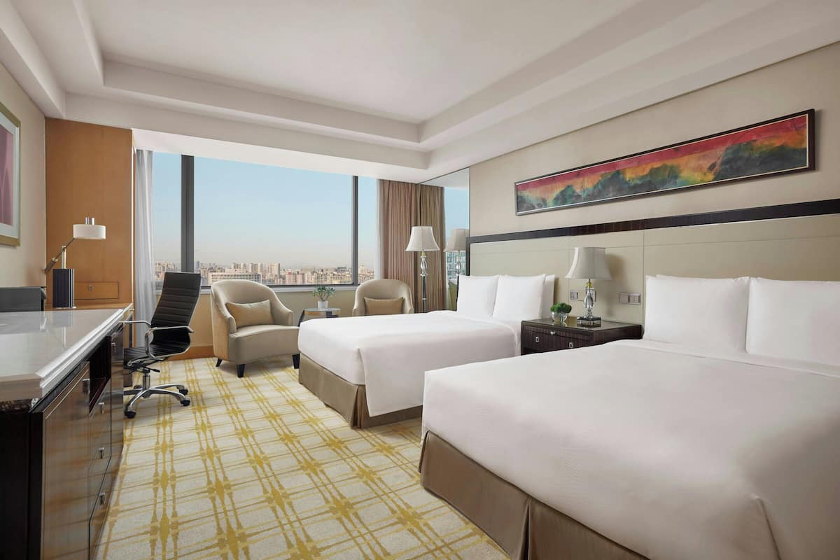A hotel room with two beds, a desk, a chair, a lamp, and a large window offering a city view.