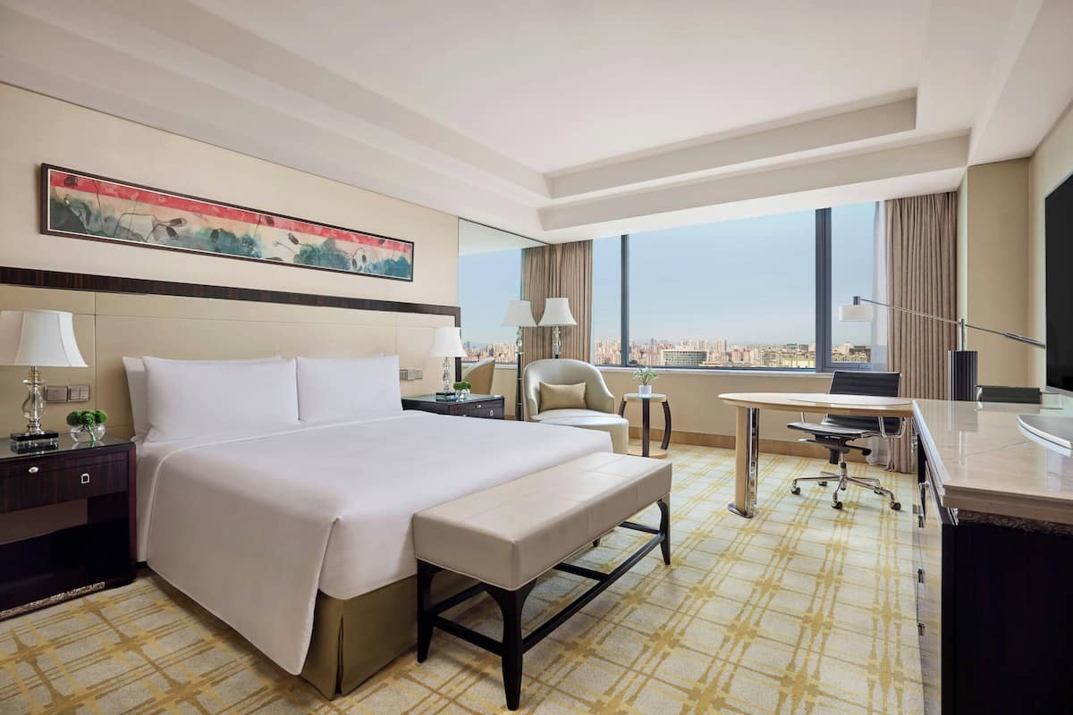 A hotel room with a large bed, a desk, a chair, a bench, a desk lamp, a bedside table, a lamp, a painting on the wall, and a view of the city through large windows.