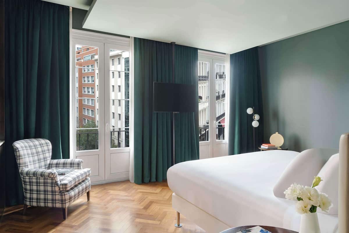 A hotel room with a large bed, a plaid armchair, a flat-screen TV mounted on the wall, and a view of a cityscape through the window.