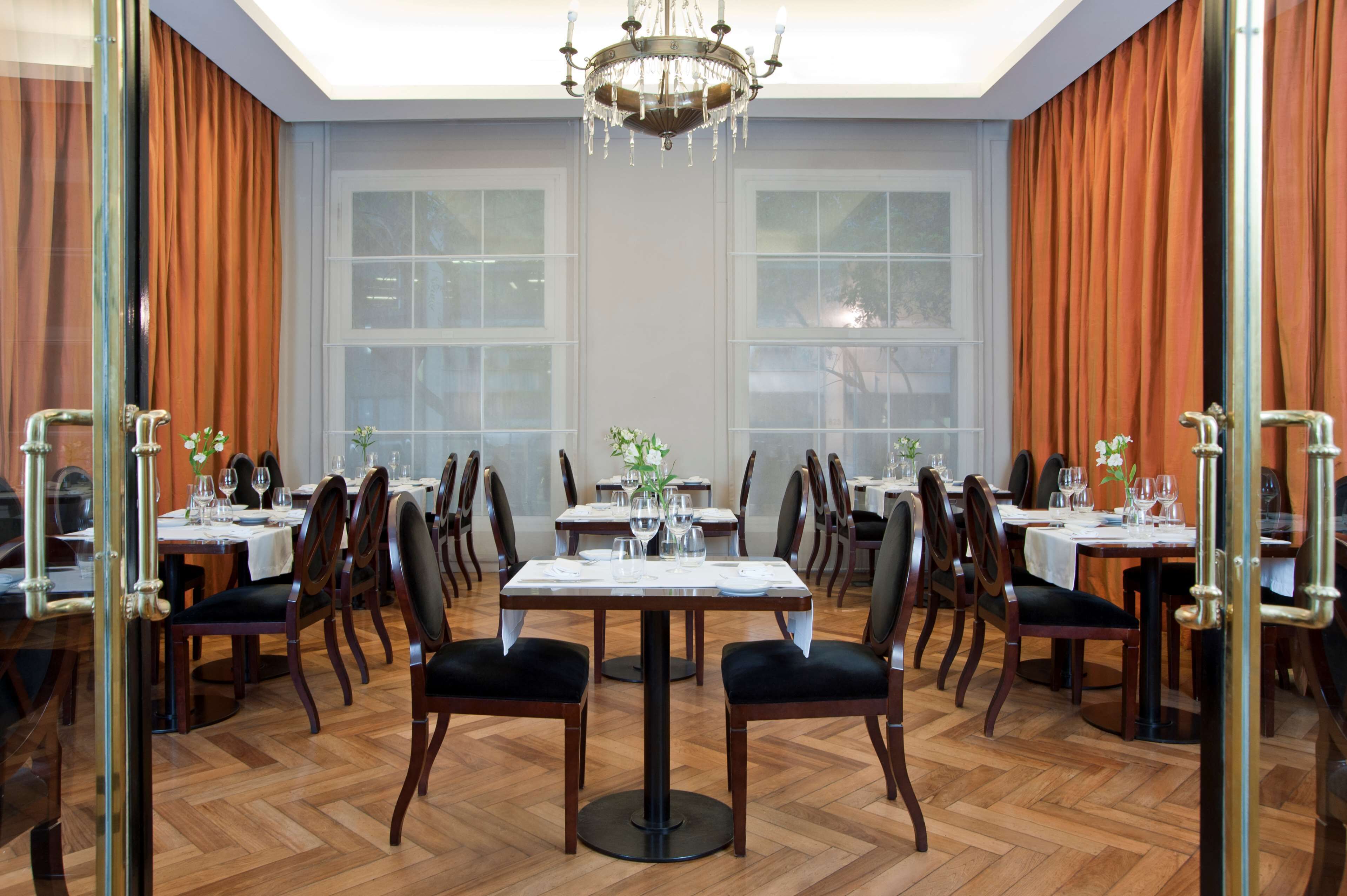 A formal dining room with multiple tables and chairs, large windows, and a chandelier.