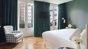 A hotel room with a large bed, a plaid armchair, a flat-screen TV, and a view of the city through large windows. - NH Collection Buenos Aires Lancaster (Buenos Aires)