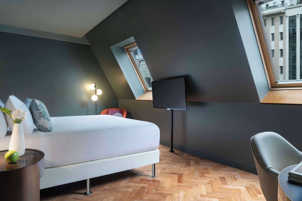 A modern bedroom with a bed, a desk, a chair, a TV, and a window with a view of buildings.