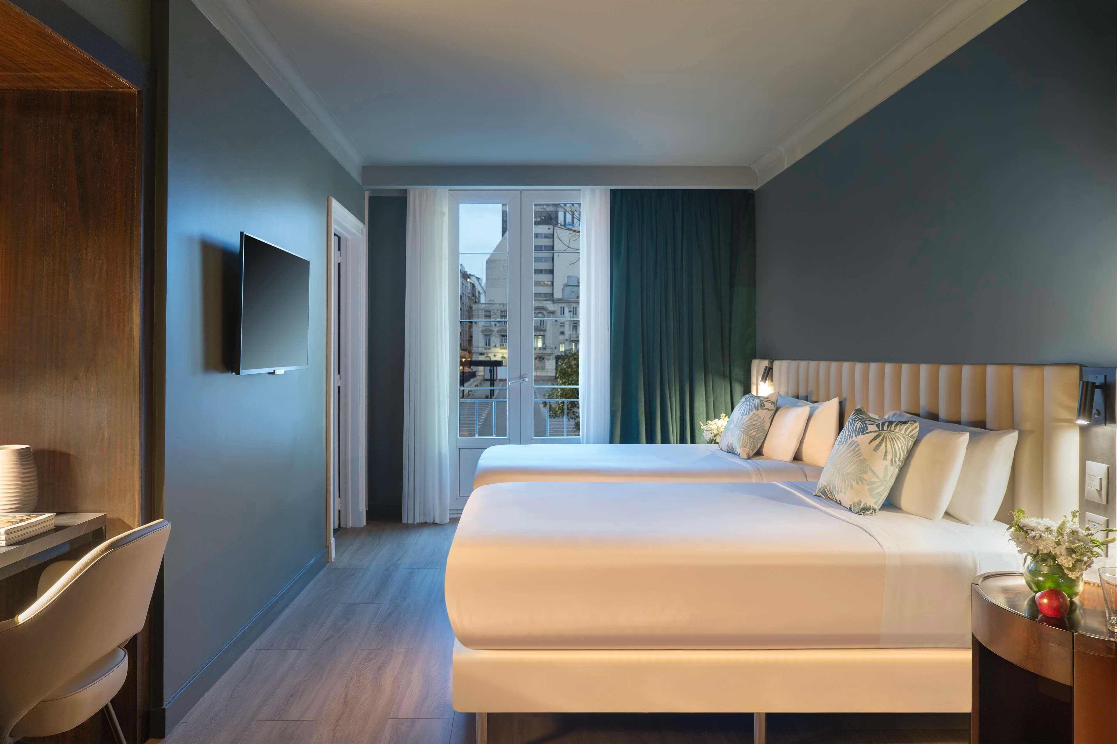 A hotel room with a bed, a TV, a desk, and a view of the city.