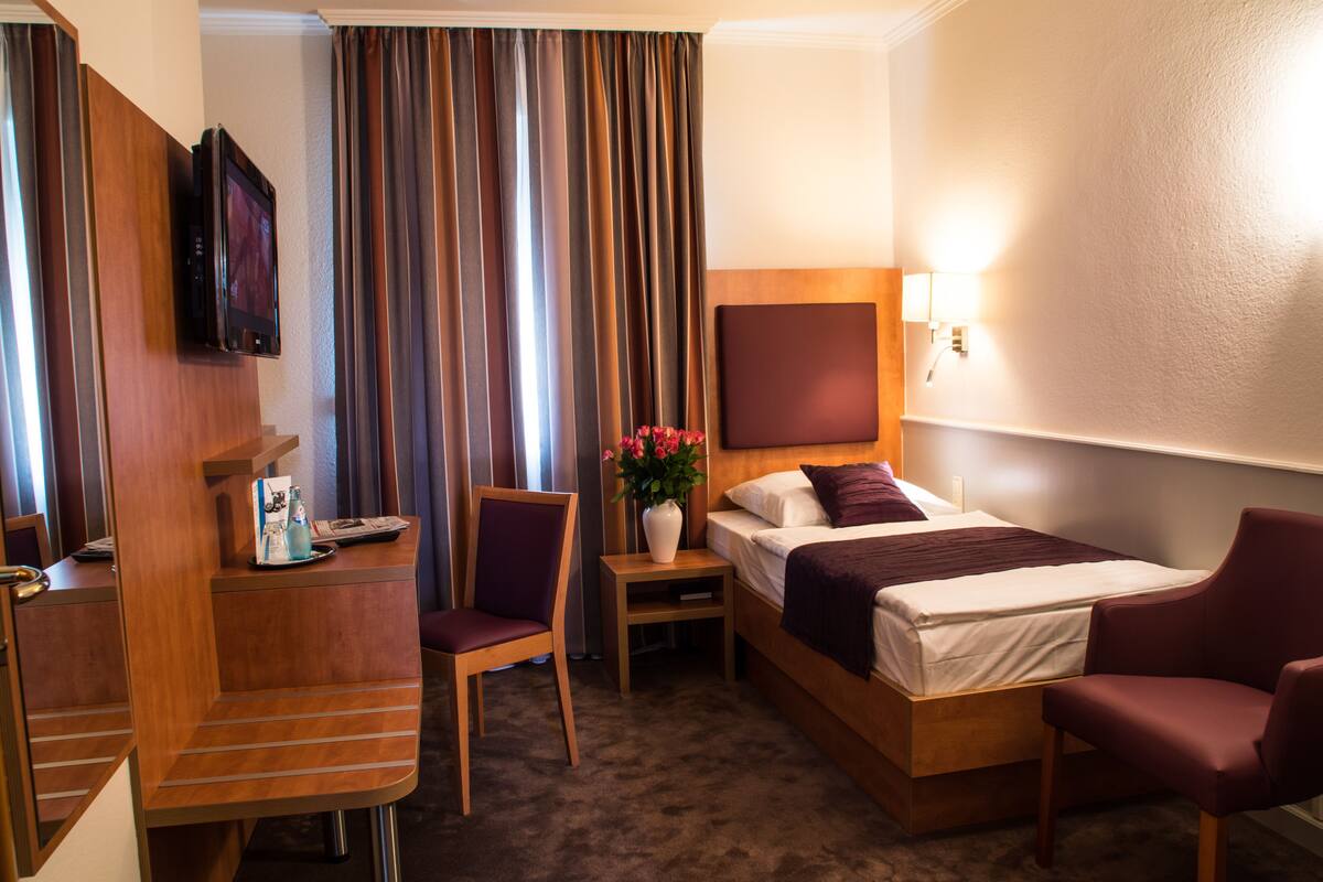 Ein Hotelzimmer mit Bett, Schreibtisch, Stuhl, Nachttisch mit Blumenvase und einem an der Wand befestigten Fernseher.