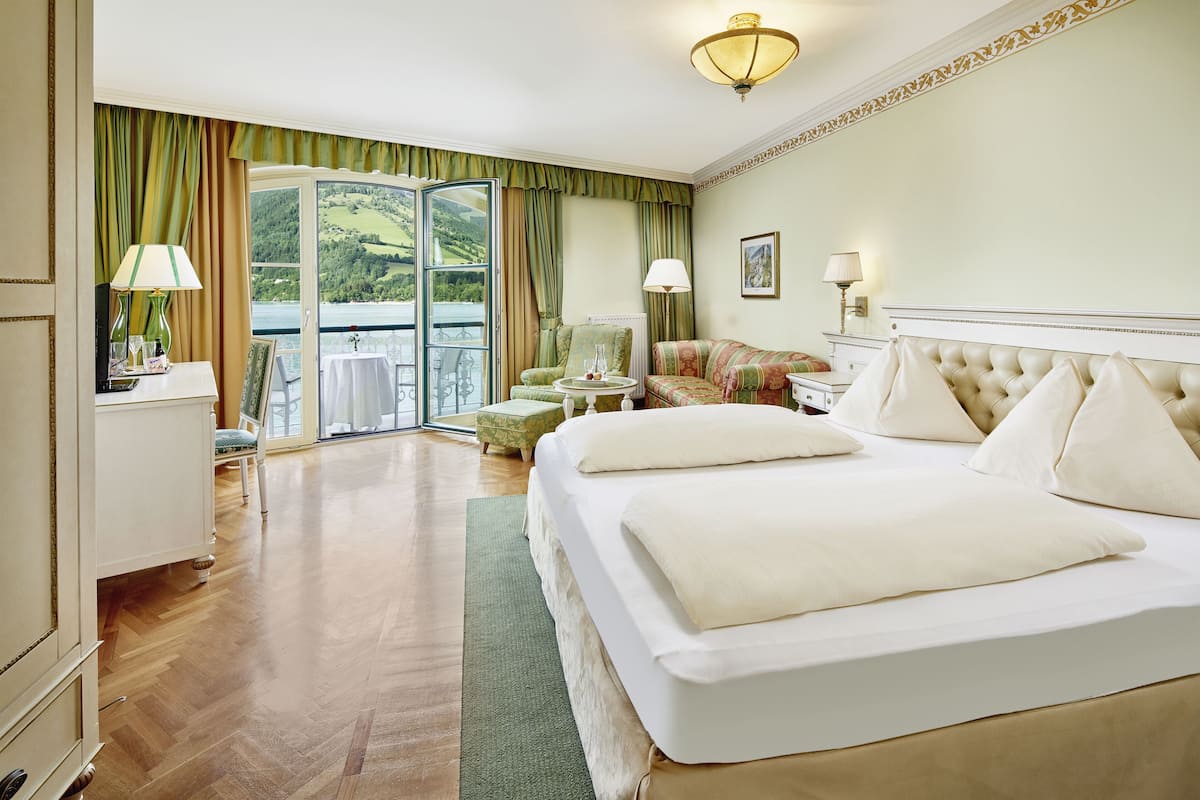 Ein Hotelzimmer mit einem großen Bett, einem Sessel, einem kleinen Tisch und Blick ins Freie.