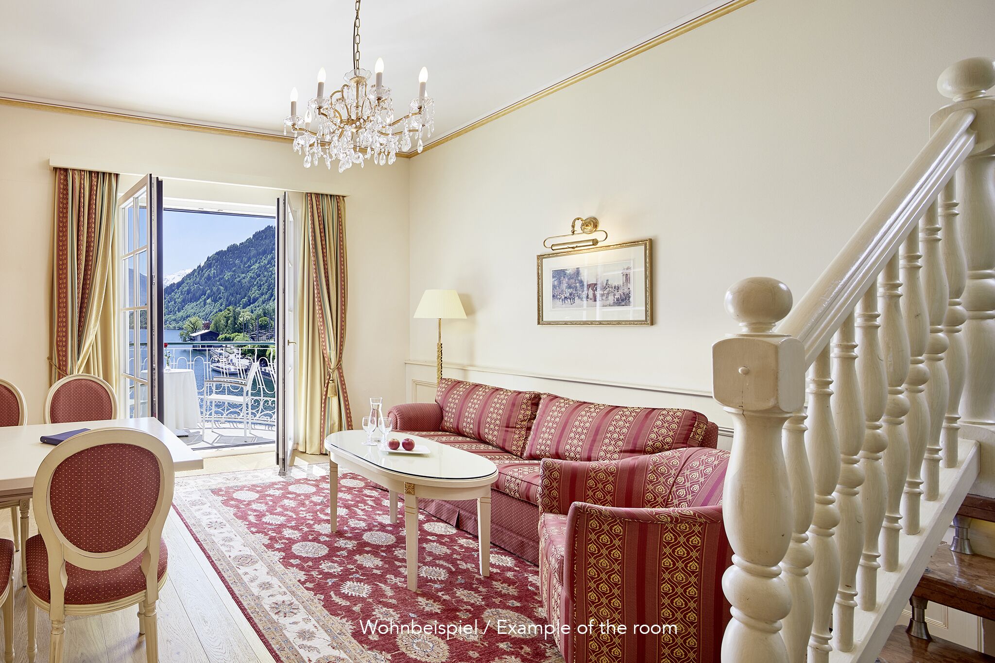 A living room with a sofa, a table, and a chandelier. There is a staircase with a railing and a view of a mountain through a window.