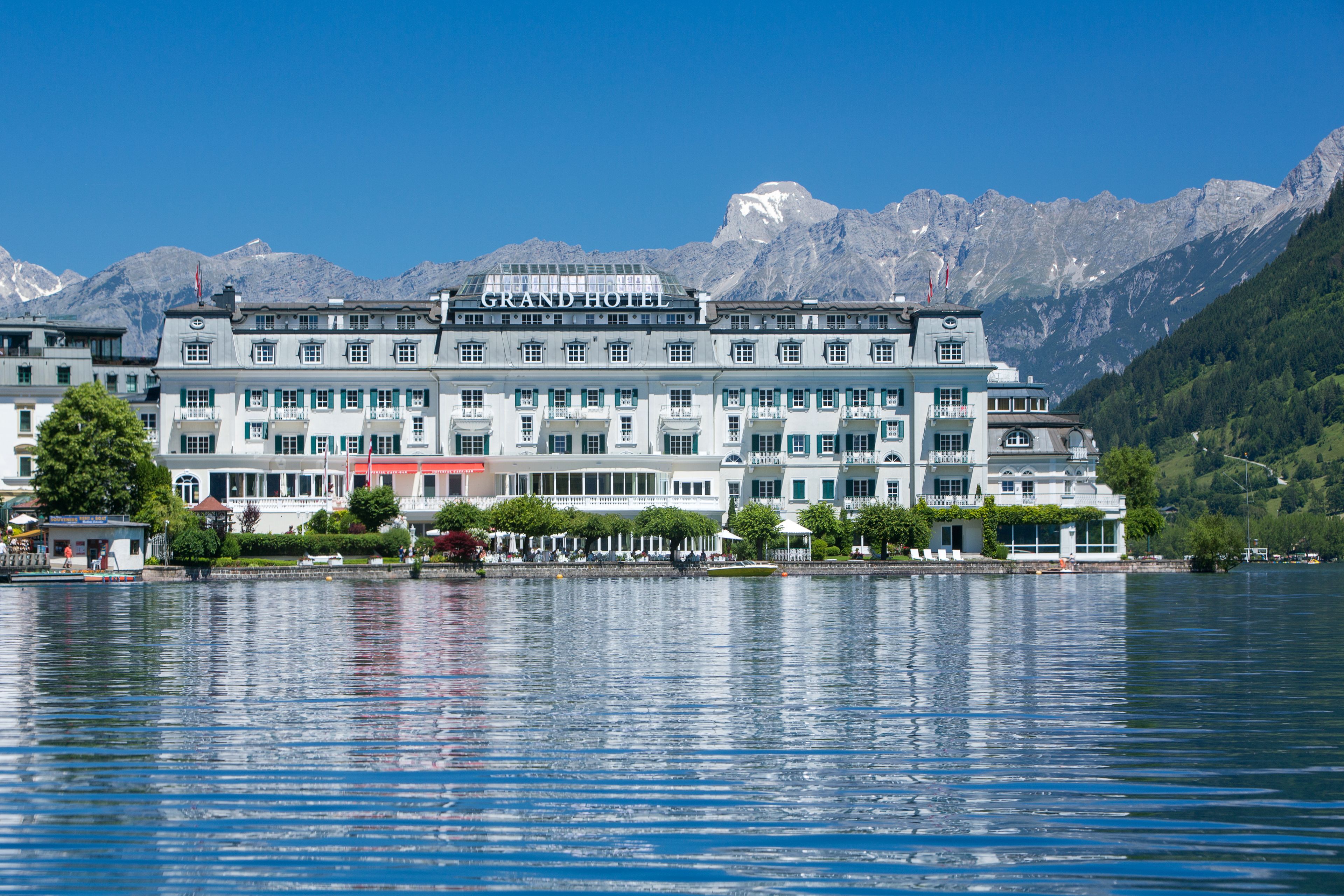 A large hotel with multiple floors and balconies, situated by a lake with mountains in the background.