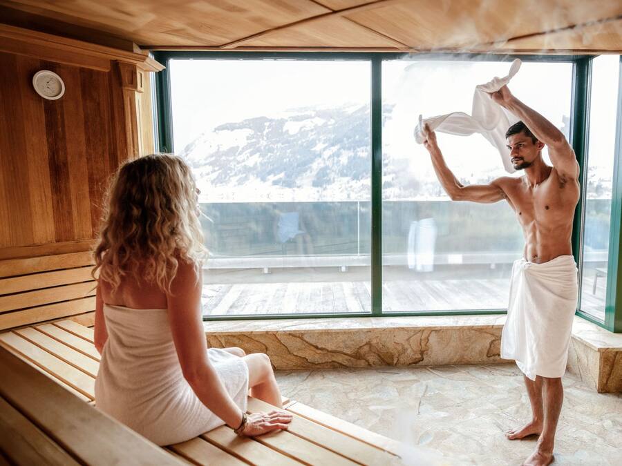 Ein Saunaraum mit Holzbänken, einem großen Fenster mit Blick auf die Berge und einer Person mit Handtuch.