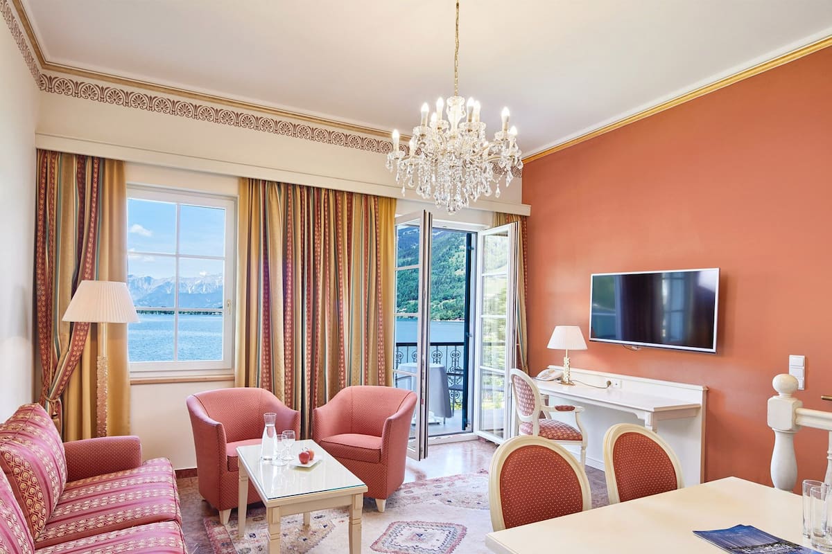 Une chambre d’hôtel dotée d’une grande fenêtre, d’un lustre, d’un téléviseur à écran plat et d’un coin repas avec une table et des chaises.