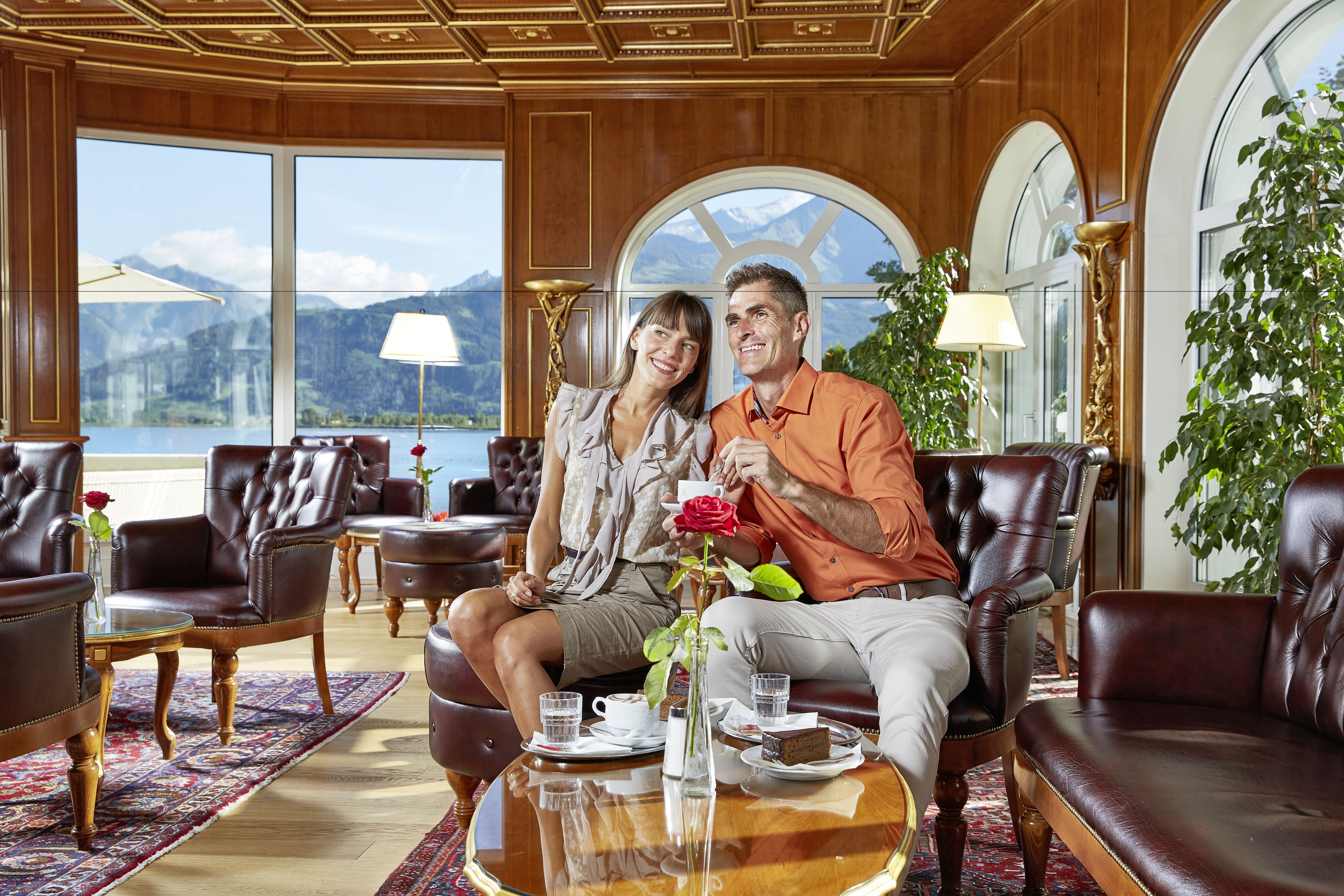 Una habitación lujosa con mobiliario de cuero, una mesa de vidrio con un jarrón y una rosa, y vistas a montañas a través de amplias ventanas.