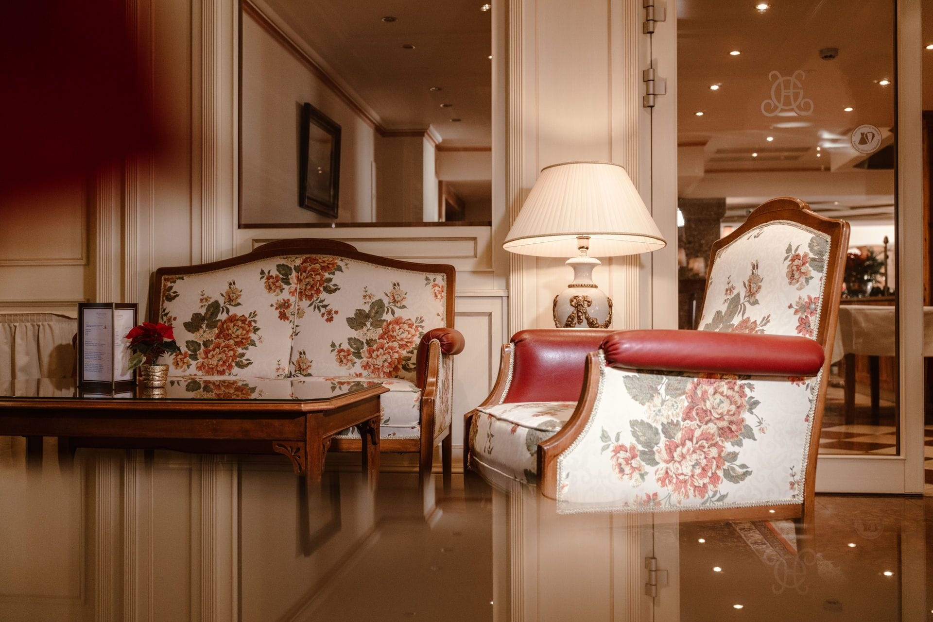 Une pièce élégante et bien éclairée, dotée d’un parquet ciré, d’un canapé recouvert d’un tissu à motifs floraux, d’un fauteuil assorti, d’une table sur laquelle se trouve un vase et d’une lampe.