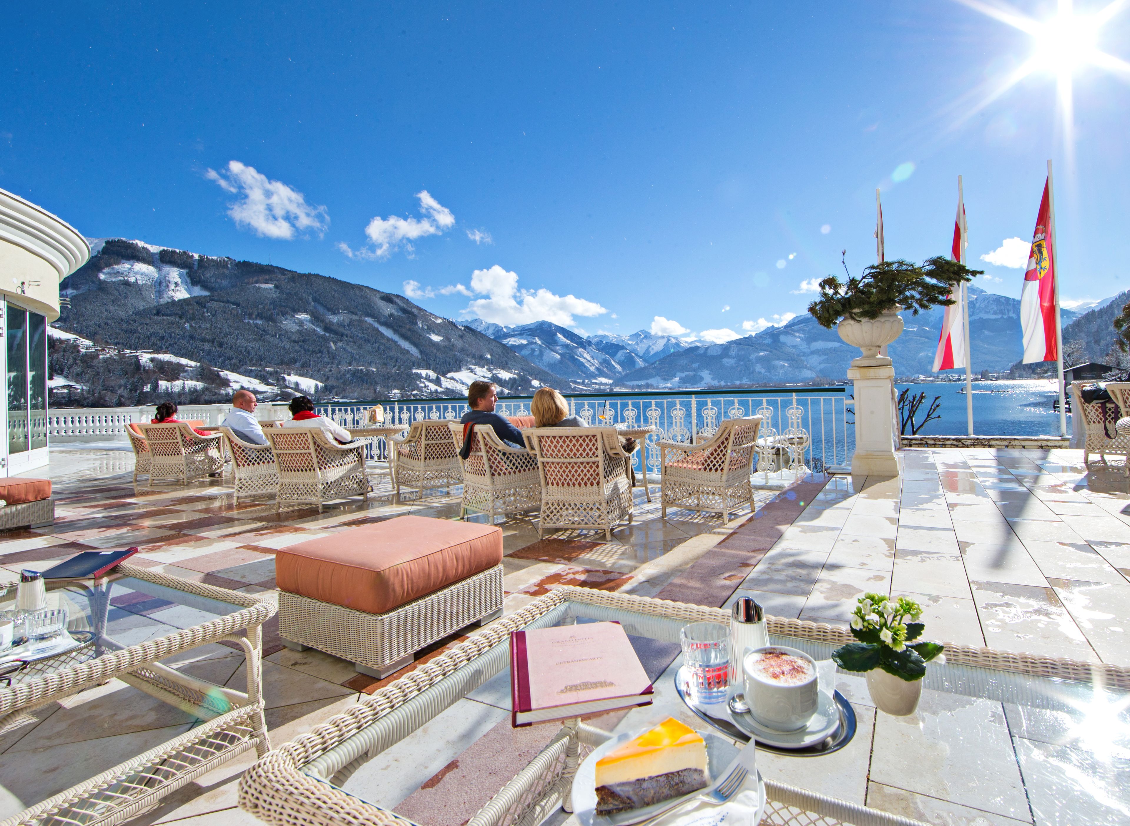 Une terrasse meublée de mobilier en osier, une table avec un livre et un verre, et une vue sur les montagnes et un lac.